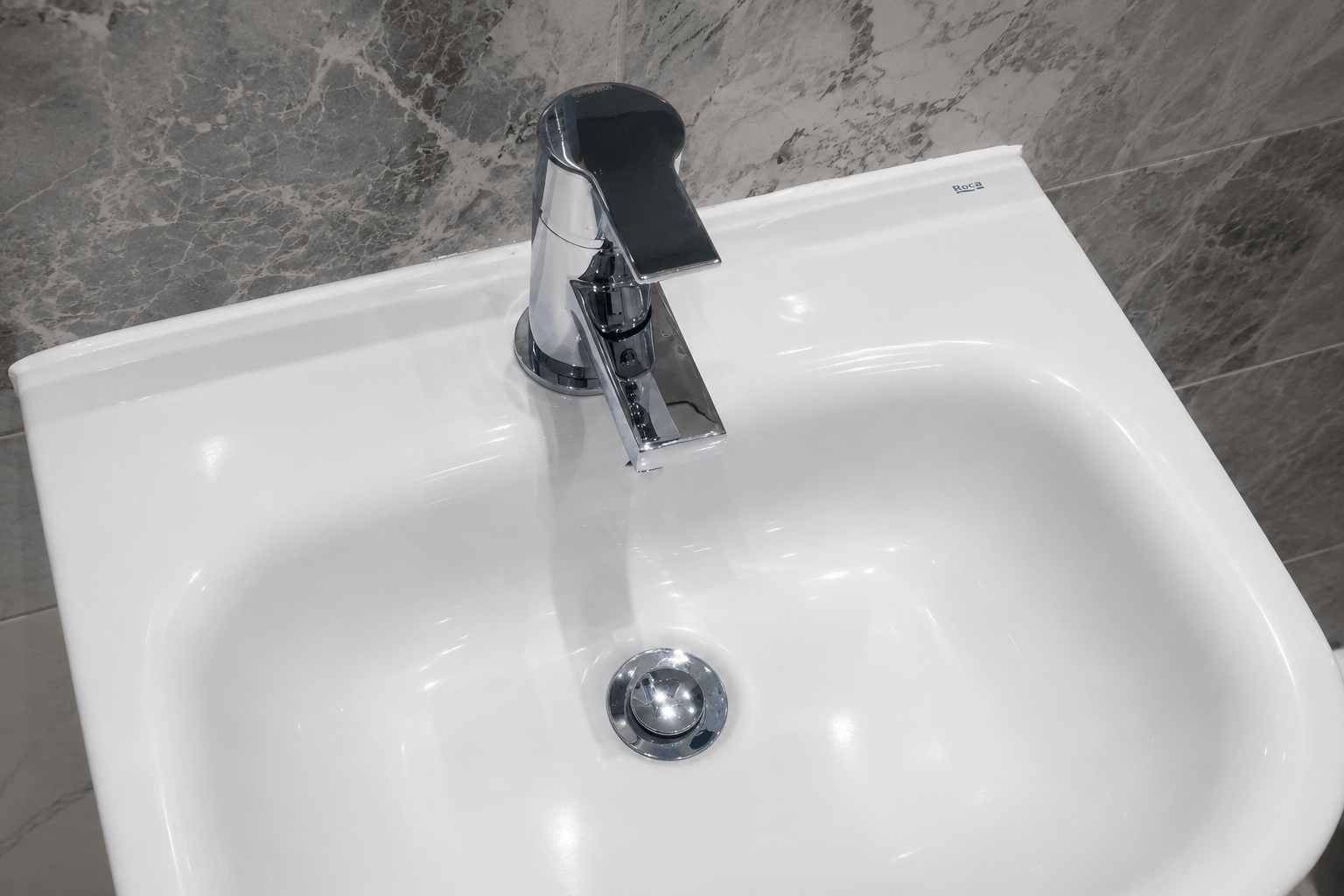 Modern white bathroom sink and chrome mixer tap after a professional deep clean, with polished surfaces and spotless grey marble-effect wall tiles.